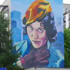 Mural przedstawiający Zofię Stryjeńską w żółtym beretce, z dłonią na piersi. Tło to błękitne, geometryczne budynki.